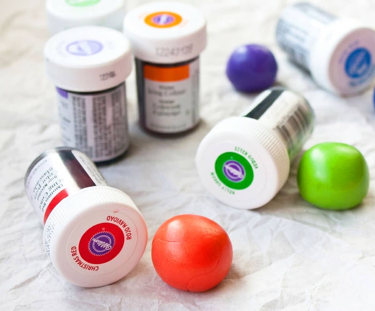 Balls of colored fondant and bottles of gel food coloring on sheet of parchment paper. 