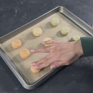 Cookie dough balls on baking tray and being flattened with hands. 
