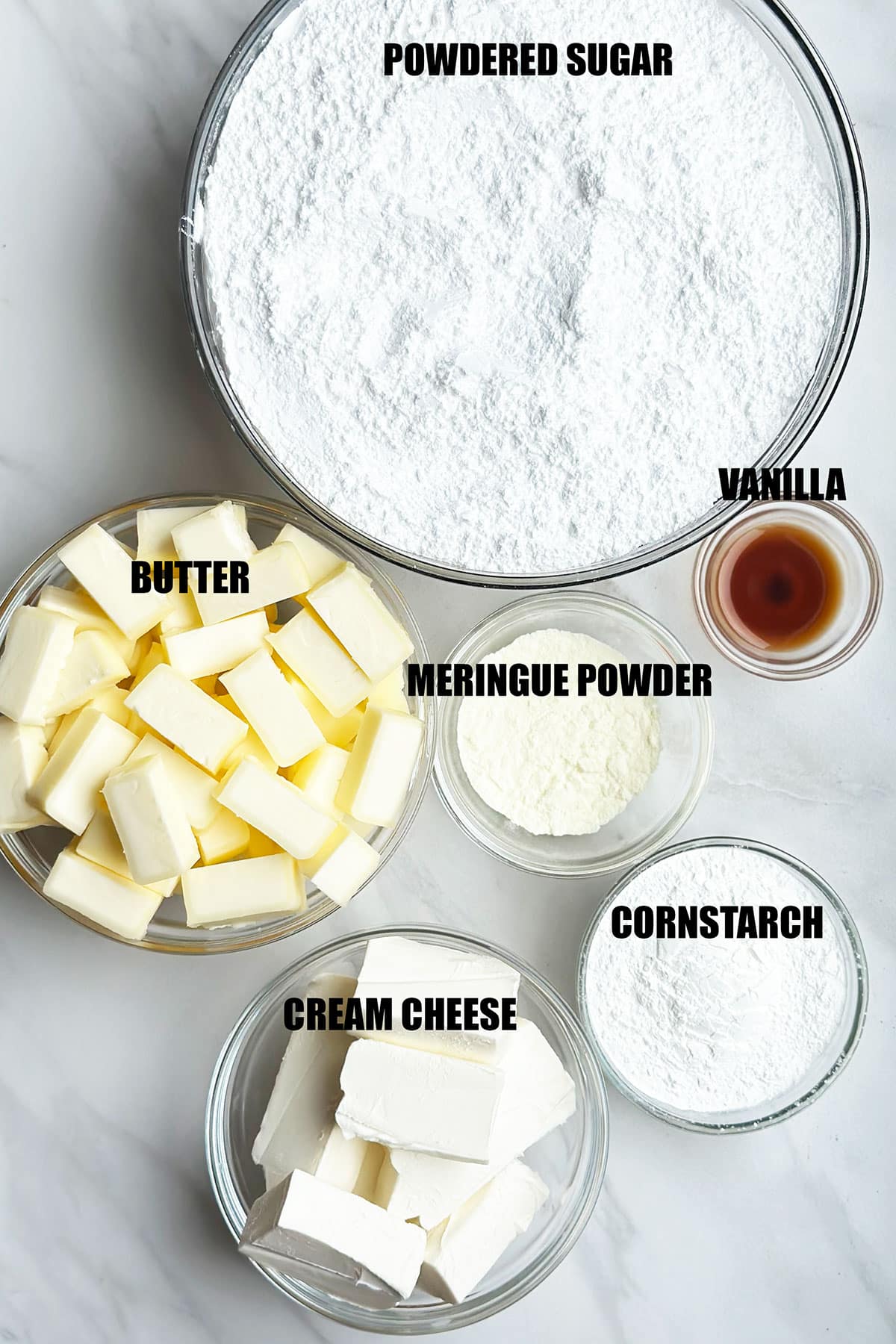 Ingredients for recipe on white marble background- powdered sugar, vanilla extract, butter, meringue powder, cornstarch, cream cheese. 