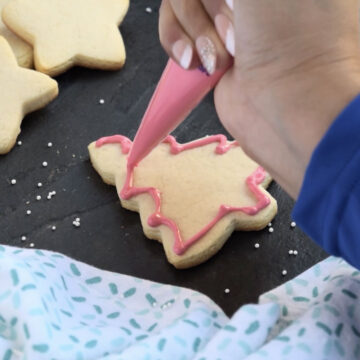 Thick pink icing being used to pipe the outline on a cut out tree cookie. 