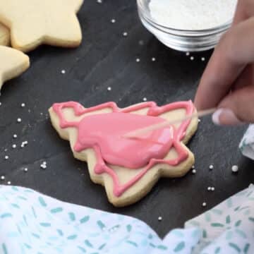 Pink flood icing being used to fill cut out tree cookie with color. 