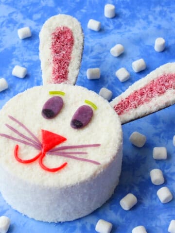 Easy Classic Easter Bunny Cake With White Cream Cheese Frosting, Candies and Coconut on Blue Background With Marshmallows.