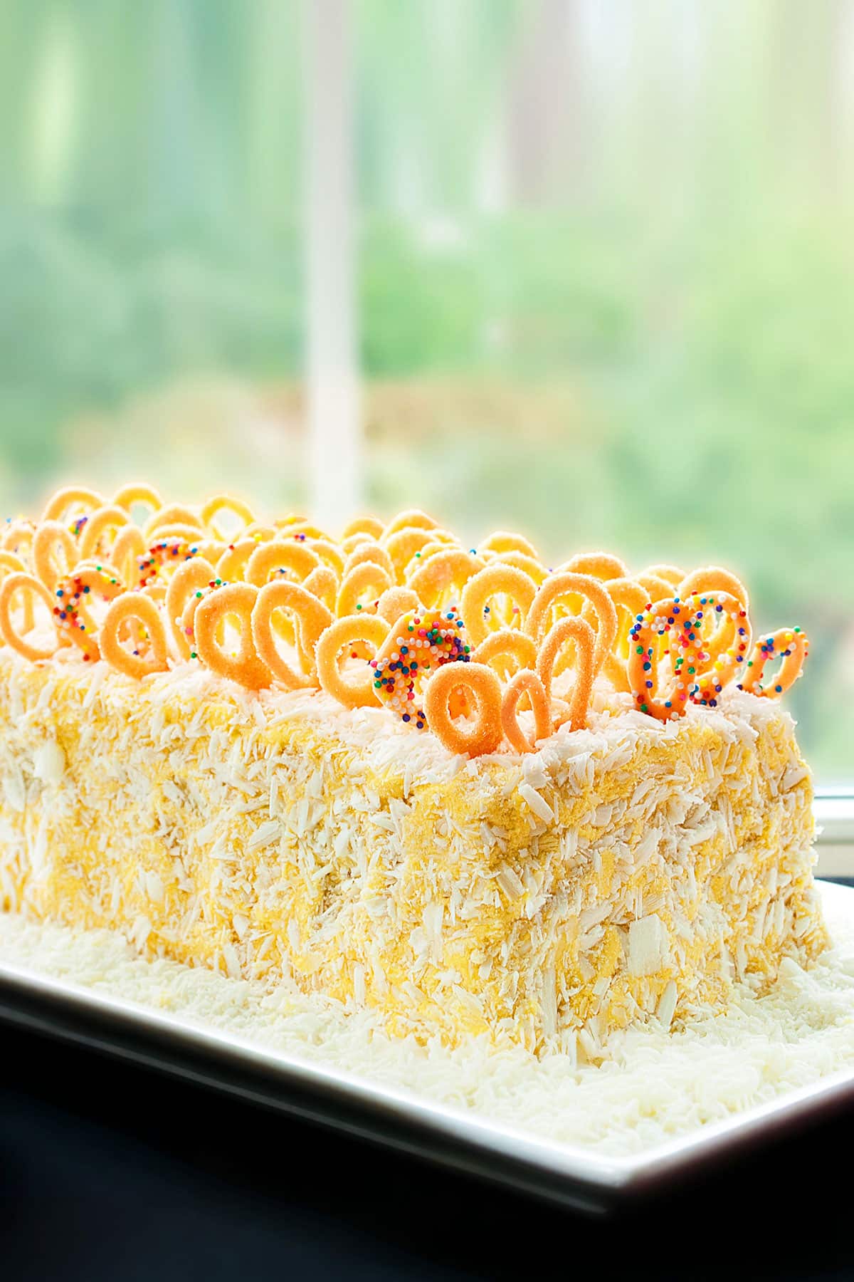 Easy Homemade Soft Mango Cake With Mango Buttercream Frosting and Chocolate Decorations on White Dish Near Window.