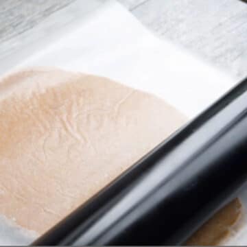 Cookie dough being rolled between two sheets of wax paper. 