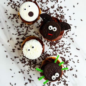 Easy Homemade Halloween Cupcakes (Mummy, Ghost, Spider, Bat) on White Marble Background With Chocolate Sprinkles Scattered Everywhere.