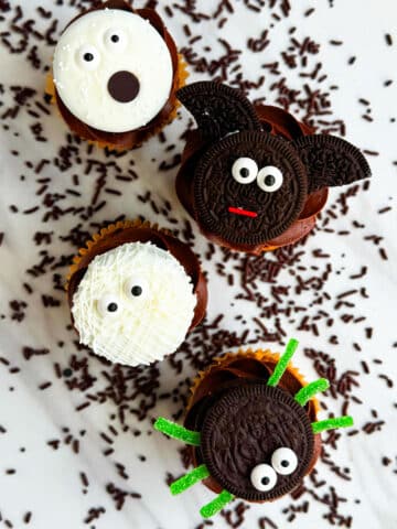 Easy Homemade Halloween Cupcakes (Mummy, Ghost, Spider, Bat) on White Marble Background With Chocolate Sprinkles Scattered Everywhere.