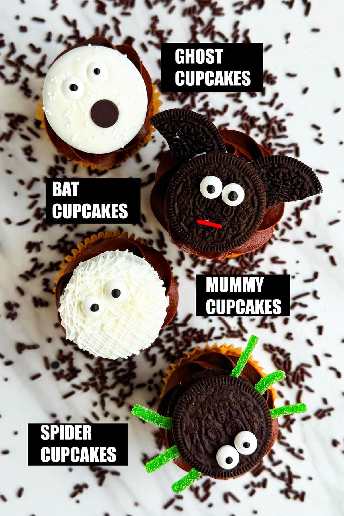 Easy Labeled Homemade Halloween Cupcakes (Mummy, Ghost, Spider, Bat) on White Marble Background With Chocolate Sprinkles Scattered Everywhere.