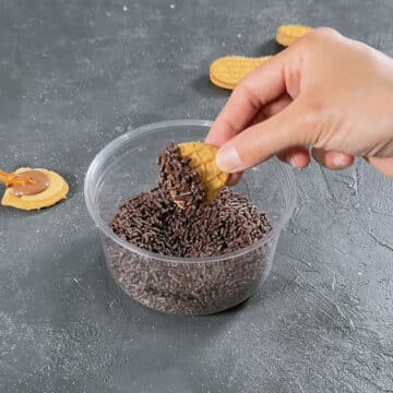 Cookie being dipped in plastic bowl of chocolate sprinkles. 