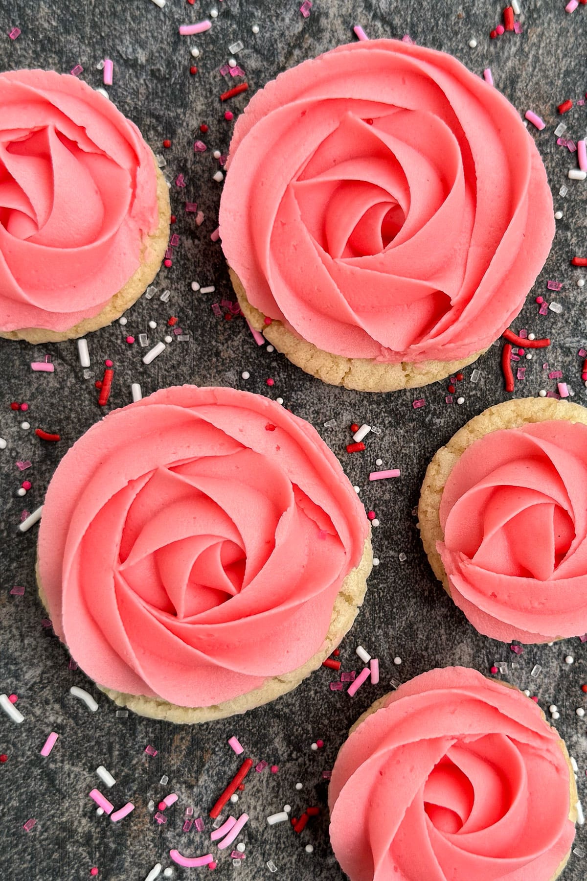 Easy rose cookies with pink buttercream frosting on rustic gray background of sprinkles. 