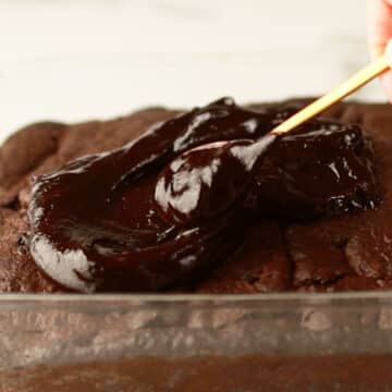 Chocolate frosting being spread on top of room temperature brownies. 