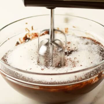 Melted chocolate mixture and sugar being mixed with a hand-held mixer. 