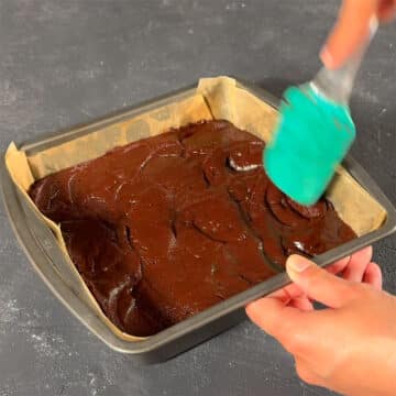 Brownie batter being spread in a lined square pan. 