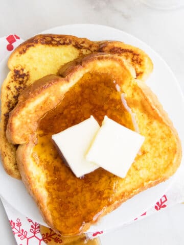 Slices of Easy Eggnog French Toast With Maple Syrup and Butter on White Dish.