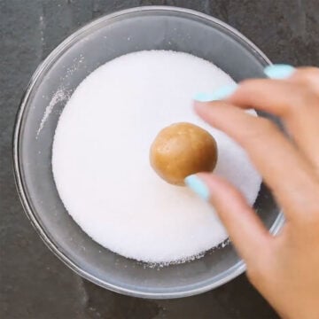 Cookie dough balls getting rolled in bowl of granulated sugar. 