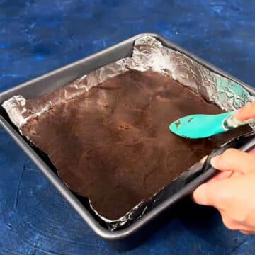 Chocolate cookie dough being spread in square baking pan with spatula. 