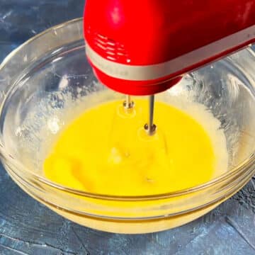 Eggs, oil and water being mixed with a hand-held mixer in a glass bowl. 