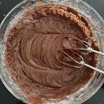 Glass bowl with chocolate icing being whipped with hand-held mixer. 