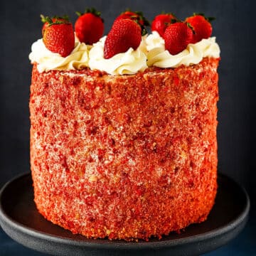 Layered Strawberry Crunch Cake With Cake Mix Box Covered in Buttercream Frosting and Crunchy Topping and Fresh Strawberries on Black Cake Stand.