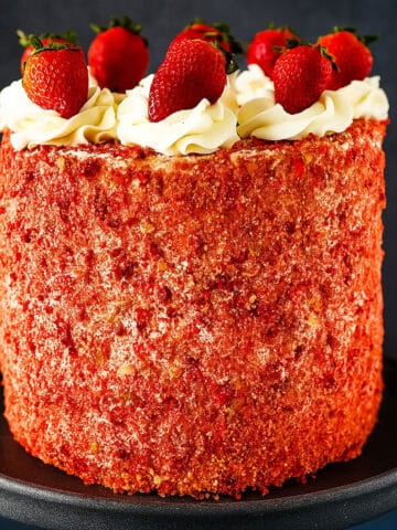 Layered Strawberry Crunch Cake With Cake Mix Box Covered in Buttercream Frosting and Crunchy Topping and Fresh Strawberries on Black Cake Stand.