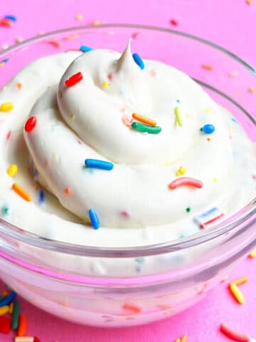 Glass Bowl of Funfetti Cake Batter Dip Topped With Colorful Sprinkles on Pink Background.