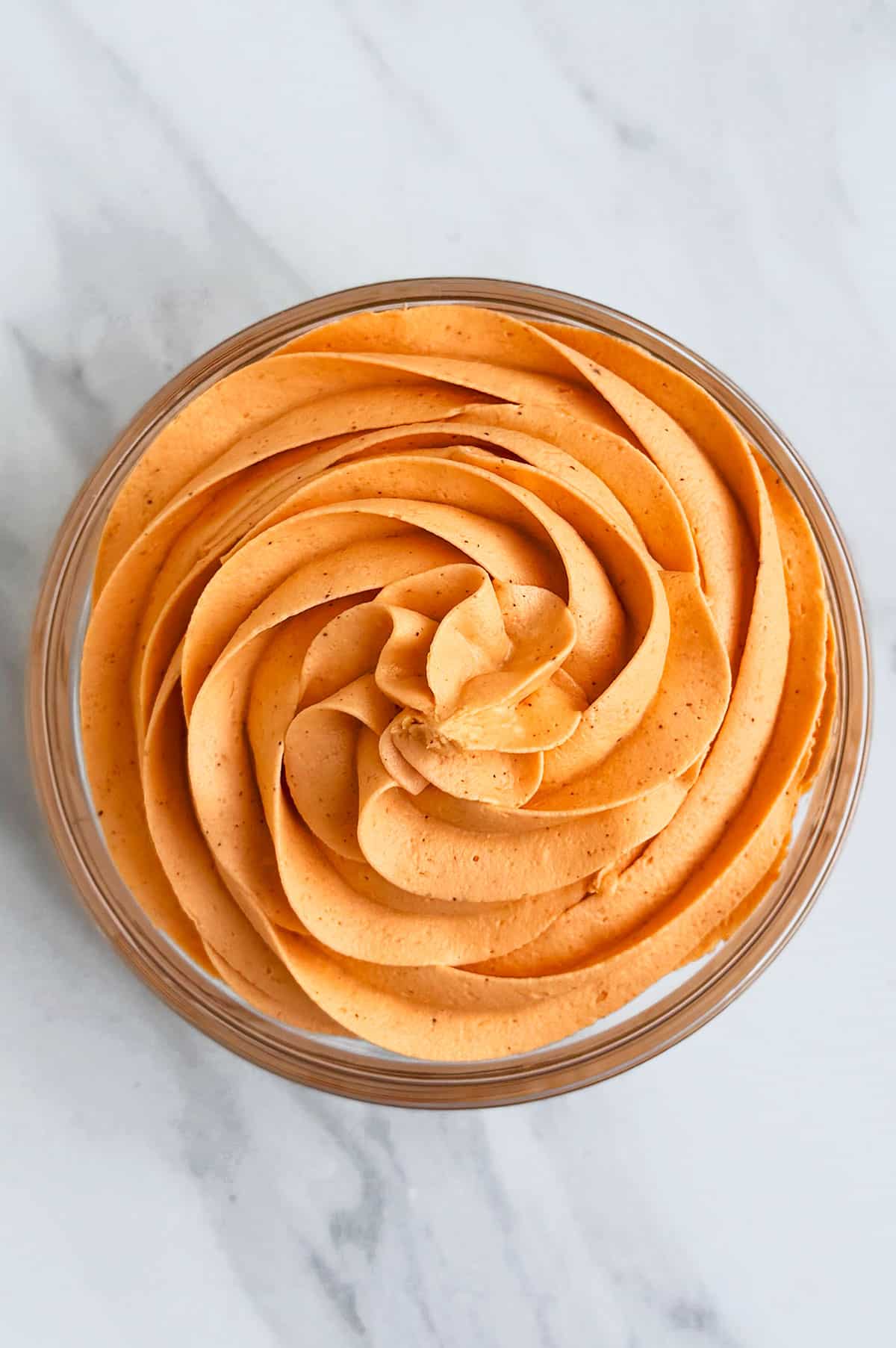 Glass bowl of spiced buttercream frosting on white marble background. 