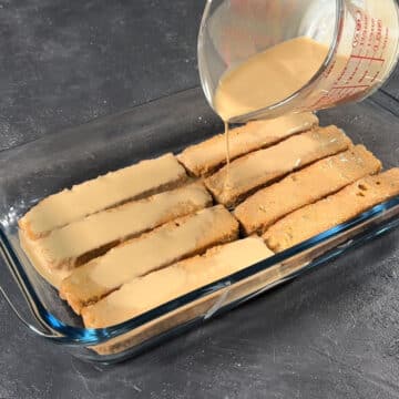 Chai soaked cake rusks arranged in a dish with more chai being poured on top. 