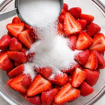 Sugar being added to sliced strawberries in glass bowl. 