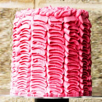 Decorated Pink Buttercream Ruffle Cake on Cake Stand with Brick Background.