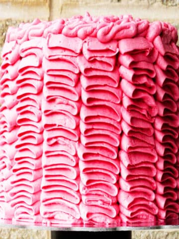 Decorated Pink Buttercream Ruffle Cake on Cake Stand with Brick Background.