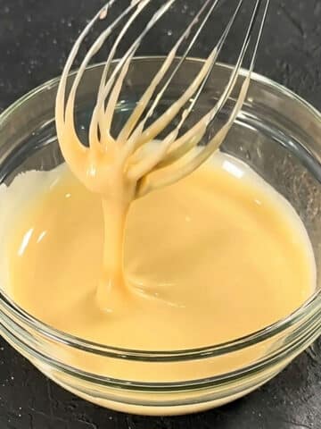 Glass Bowl Filled With Easy Maple Glaze Drizzle Icing and Stainless Steel Whisk on Rustic Gray Background.