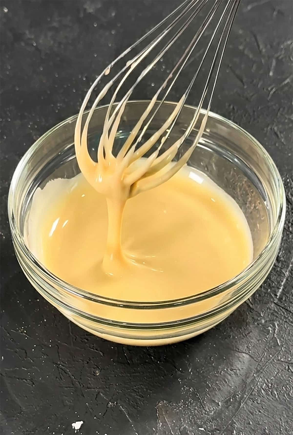 Glass Bowl Filled With Easy Maple Glaze For Cake and Desserts and Stainless Steel Whisk on Rustic Gray Background. 
