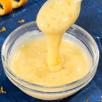 Spoonful of easy orange glaze drizzle icing for cake being poured in a glass bowl.