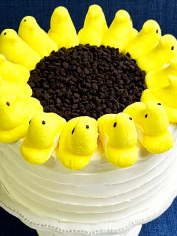 Easy Simple Sunflower Cake Decorated With Buttercream Frosting, Peeps Marshmallows and Chocolate Chips on White Cake Stand.