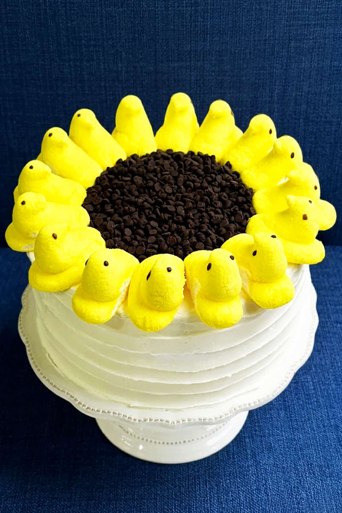 Easy Homemade Sunflower Cake Decorated With Buttercream Frosting, Yellow Peeps Marshmallows and Chocolate Chips on White Cake Stand. 