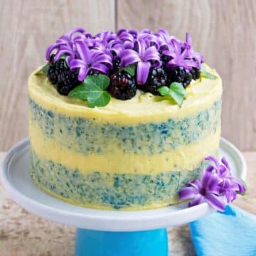 Rustic half dressed cake on stand with yellow frosting, blackberries and purple flowers.