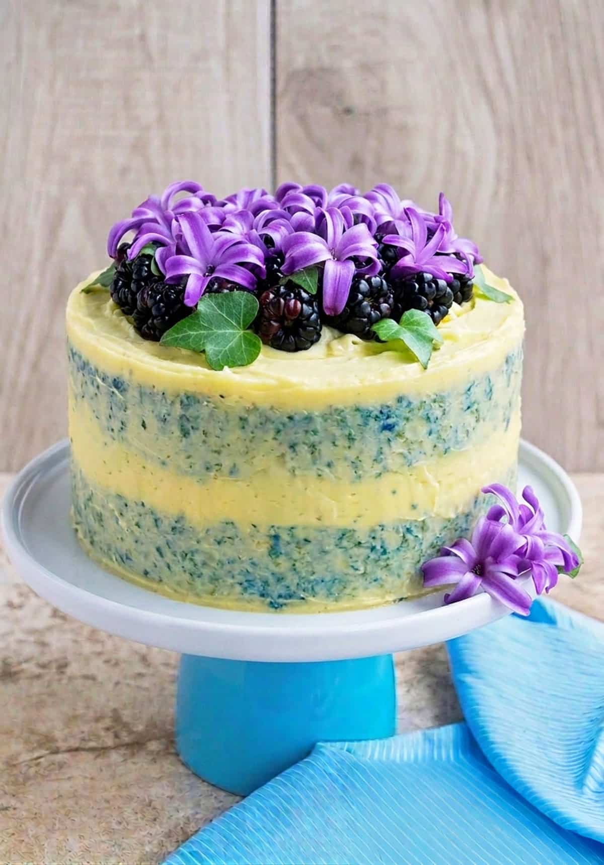 Semi naked cake with flowers and blackberries on white stand.